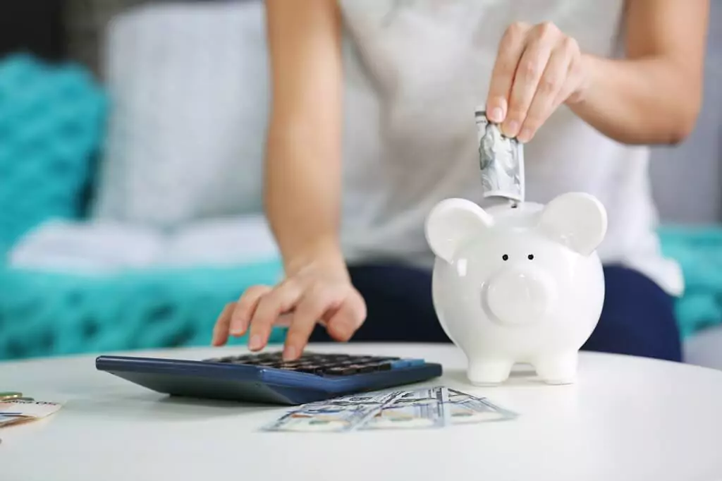 mulher colocando nota em cofrinho de porco branco, enquanto digita em calculadora