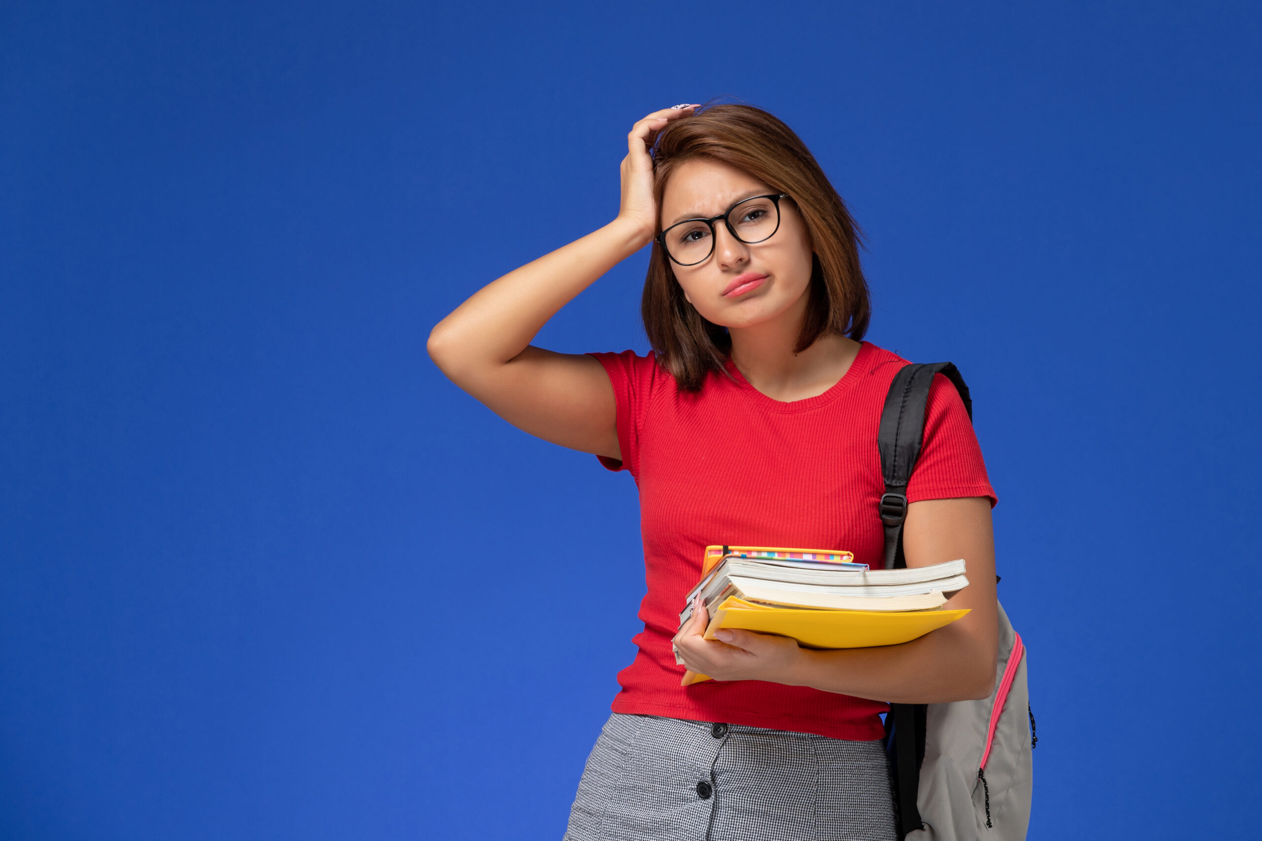 Estudante de camisa vermelha e mochila, segurando livros e pastas, com expressão preocupada