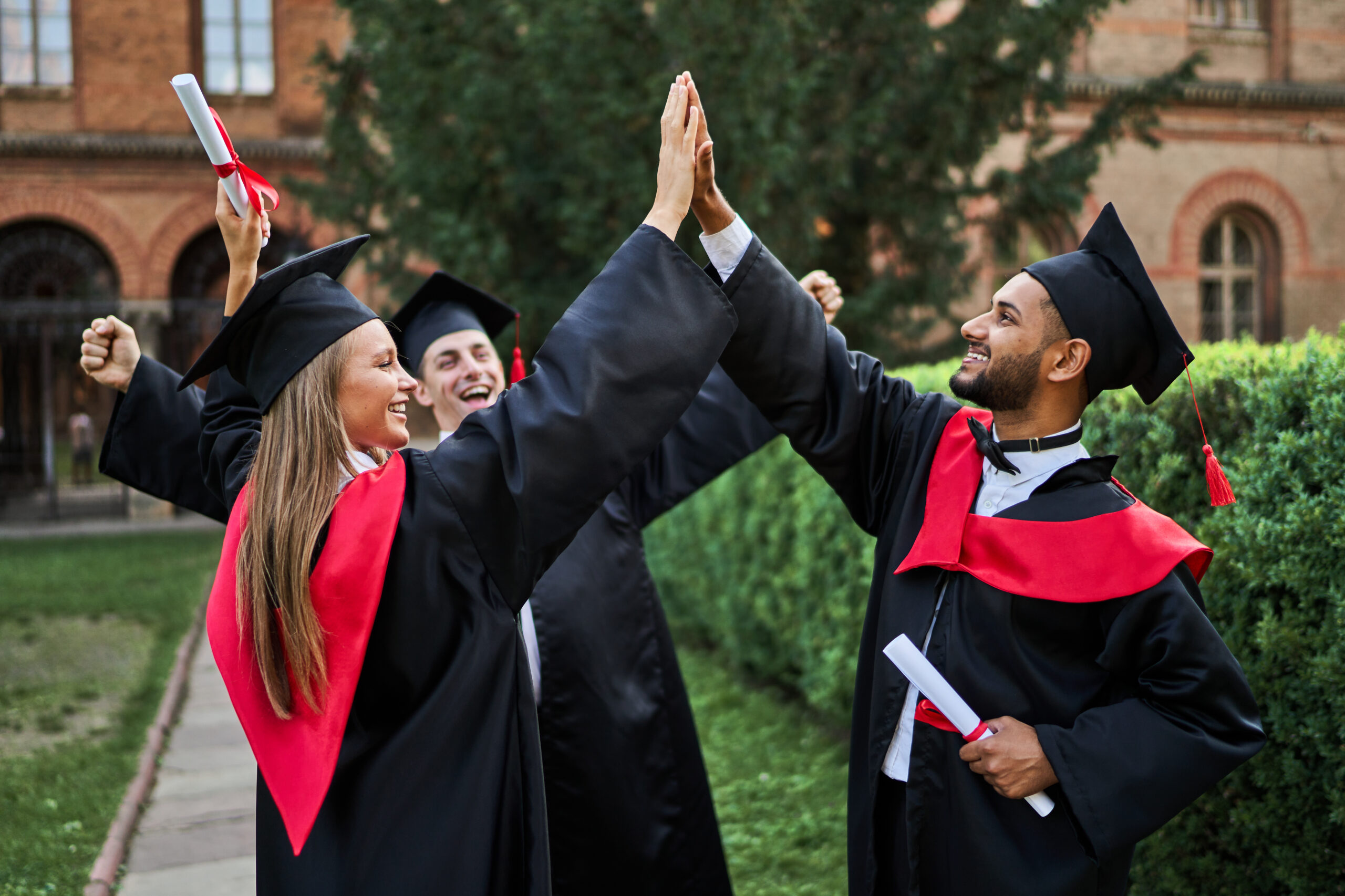 Três amigos recém-graduados, felizes, cumprimentando-se no campus da universidade, vestindo becas de formatura e segurando diplomas.